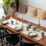 A large teak dining table beautifully decorated with candles, ceramic plates, and wine glasses, with three black dining chairs on one side and a bench on the other