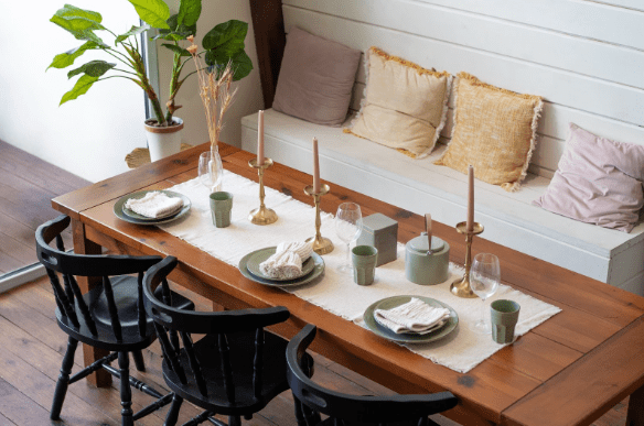 A large teak dining table beautifully decorated with candles, ceramic plates, and wine glasses, with three black dining chairs on one side and a bench on the other